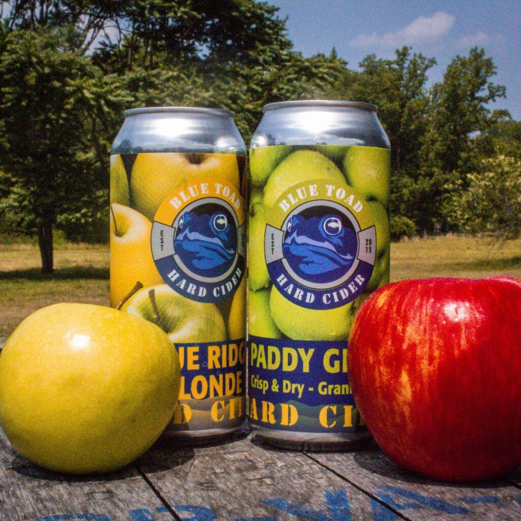 cans of cider next to apples with orchard behind